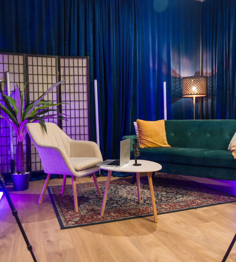 Modern content creation studio equipped for podcast recording or social media video production, featuring cameras on tripods, a microphone, laptop, sofa, armchair, and blue velvet curtains